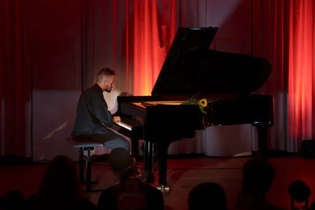 Mann spielt Klavier auf Bühne mit rotem Vorhang.