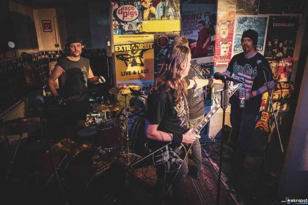 Rockband spielt live in einem Poster-geschmückten Raum.