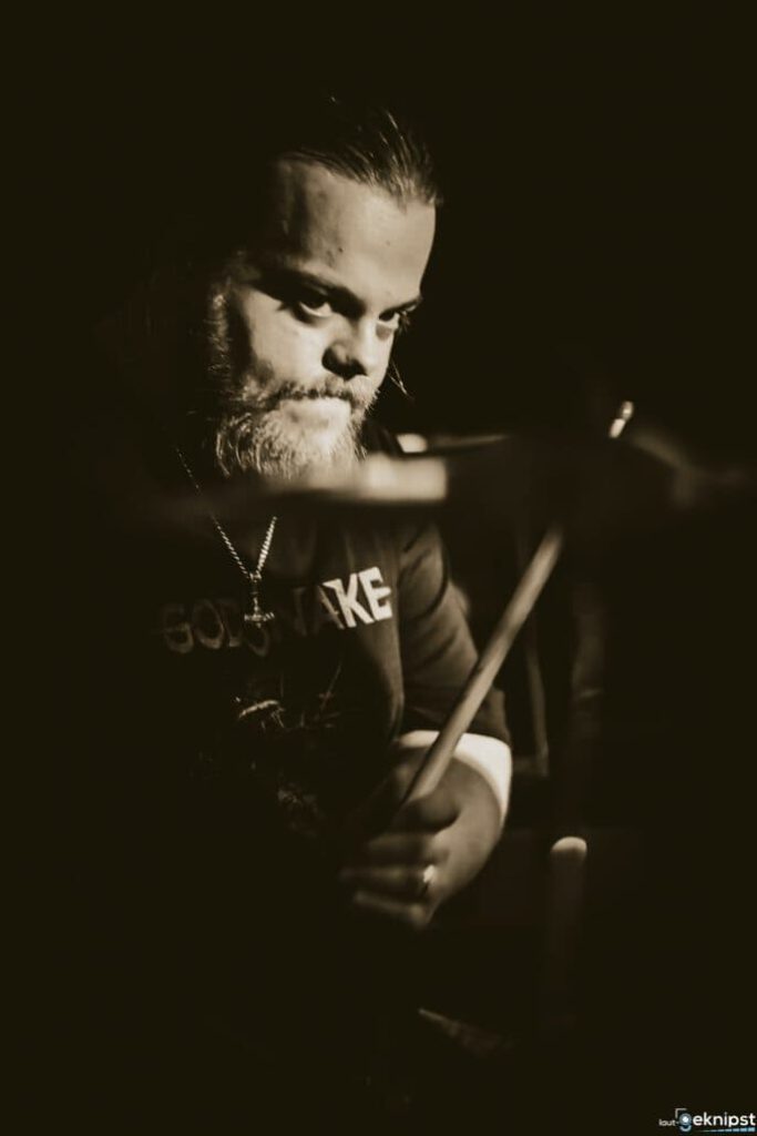 Schlagzeuger mit Bart spielt in dunklem Raum.