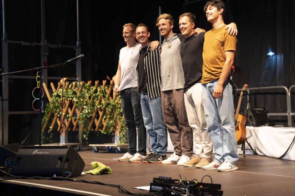 Fünf Männer lächeln auf Bühne mit Mikrofon und Gitarre.