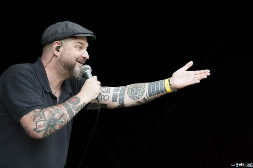 Sänger mit Tattoos spricht ins Mikrofon auf Bühne.