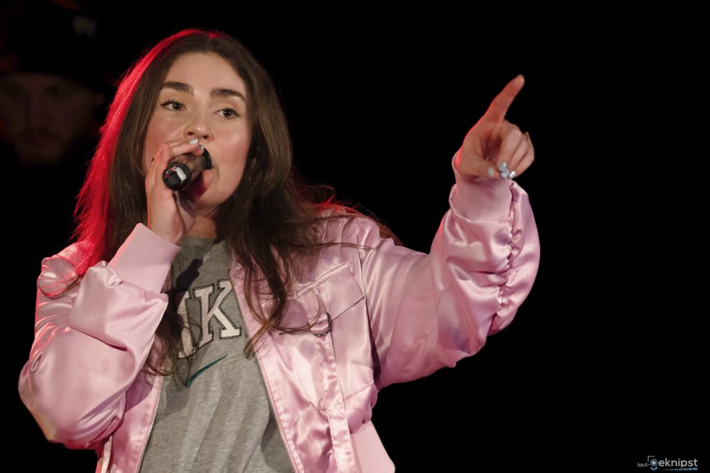 Sängerin mit Mikrofon in rosa Jacke auf Bühne.