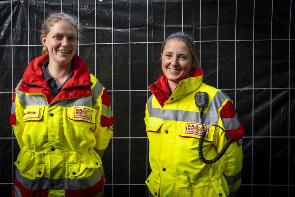 Zwei Rettungssanitäterinnen lächeln vor dunklem Hintergrund.
