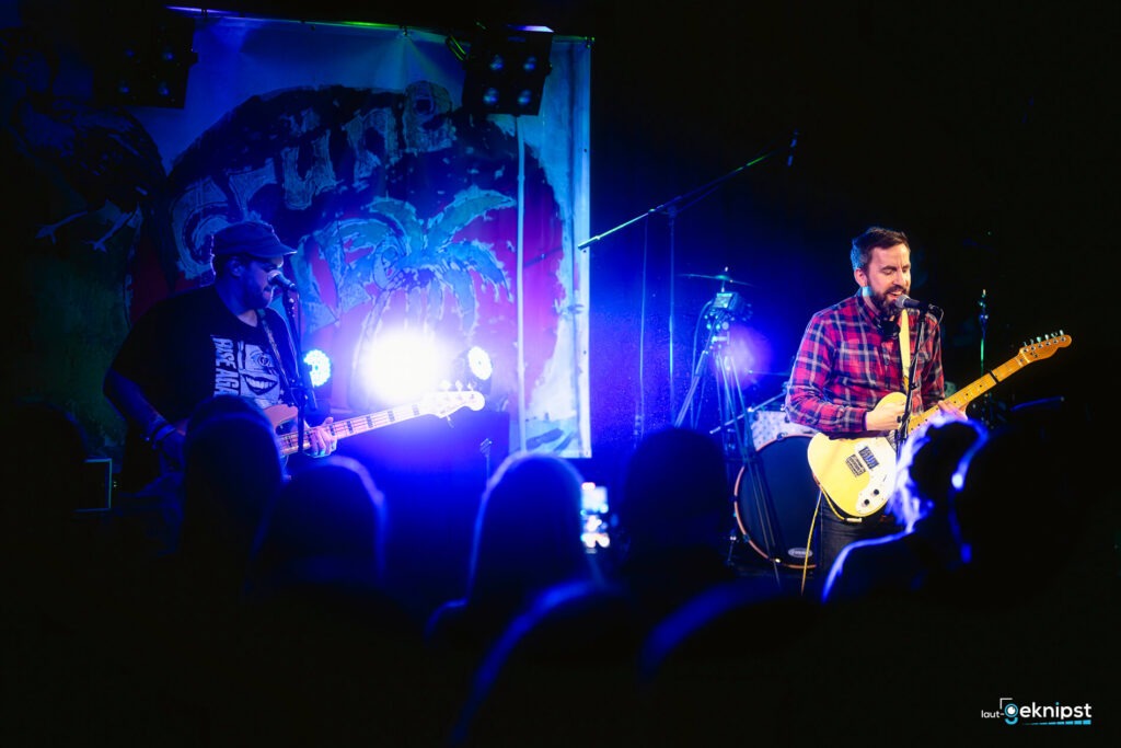 Band spielt auf Bühne mit Lichtern und Fans im Publikum.