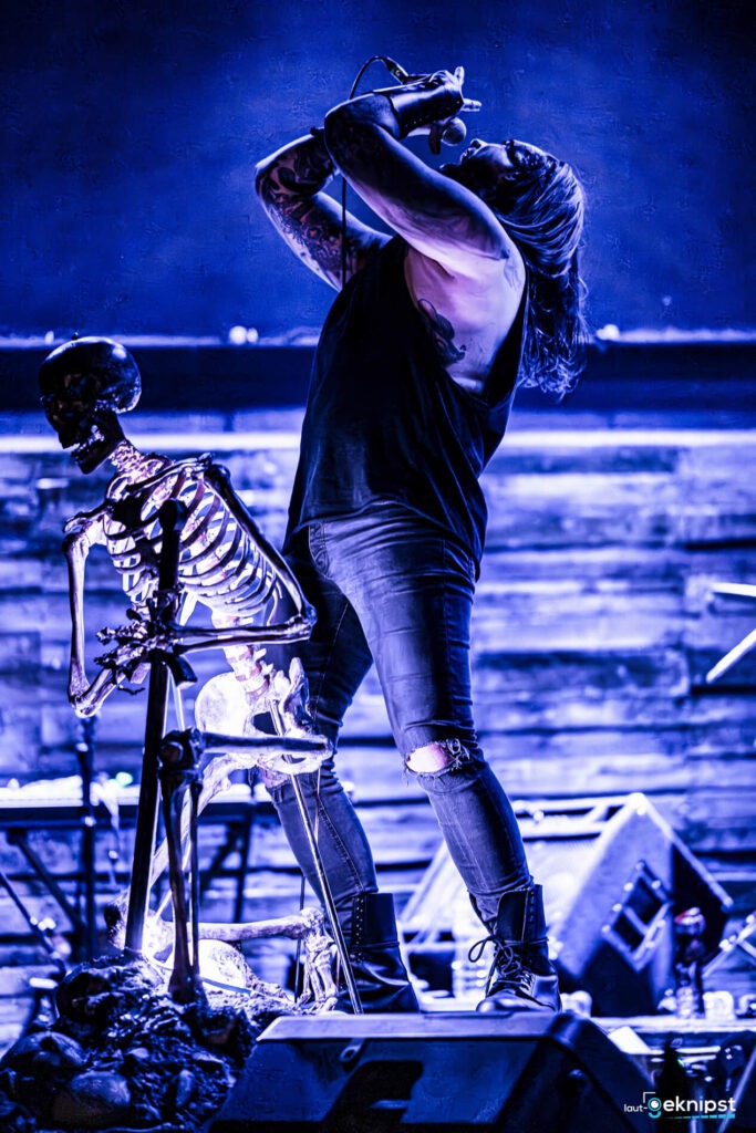 Rockmusiker singt neben Skelett auf Bühne.