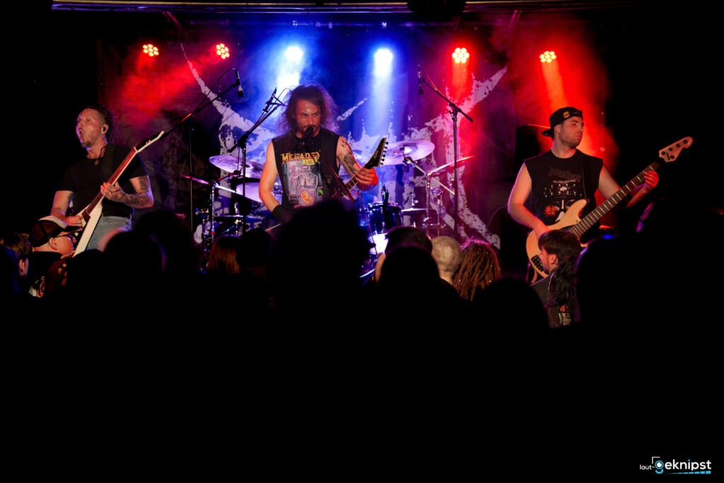 Metalband spielt live auf Bühne mit bunten Lichtern.
