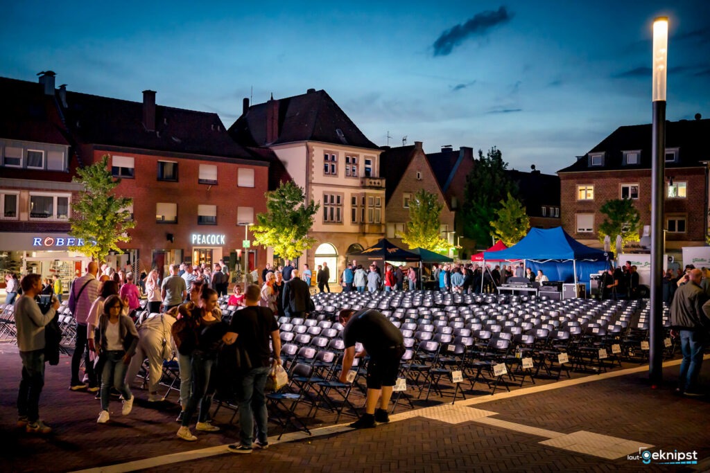 Menschen versammeln sich auf einem Platz mit leeren Stühlen.