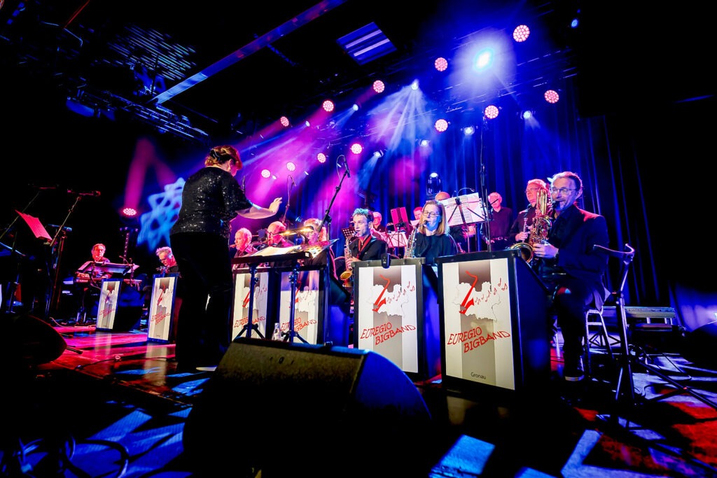 Bigband-Auftritt mit Dirigentin und Musikern auf Bühne.