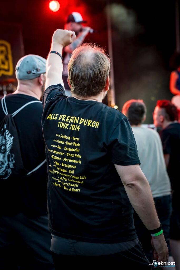 Person auf Konzert mit Tour-Shirt, Arm erhoben.