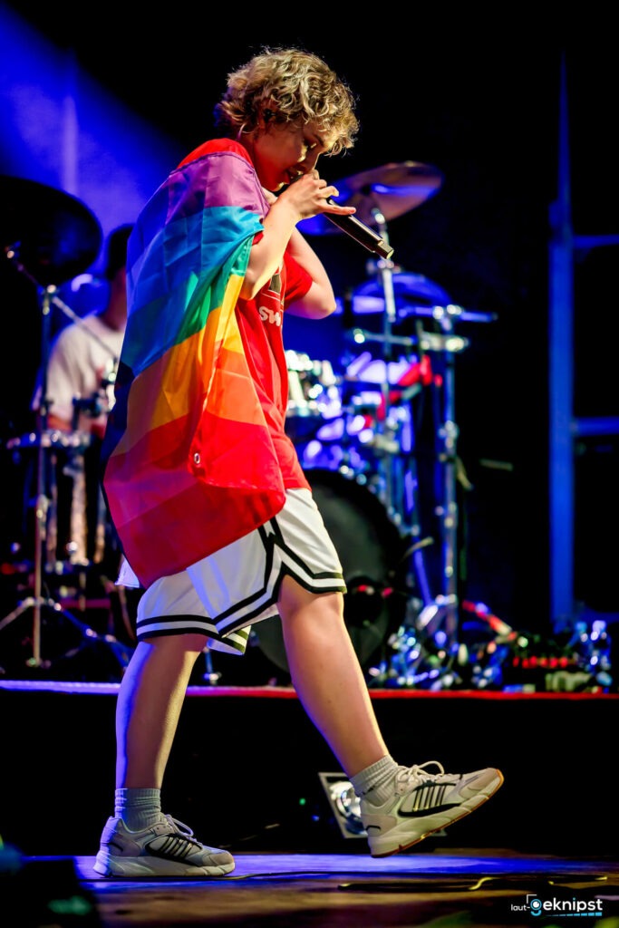 Person singt auf Bühne mit Regenbogenflagge.