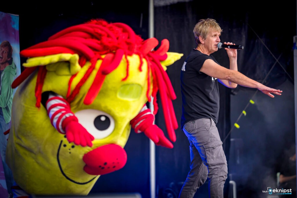 Person singt auf Bühne, begleitet von gelbem Maskottchen