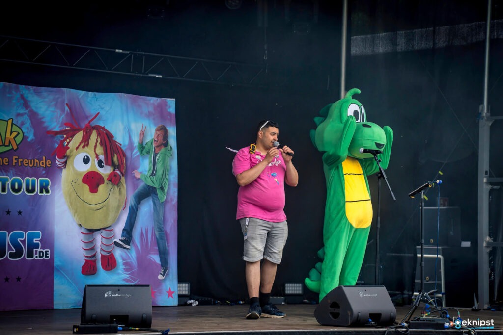 Mann und Maskottchen auf Bühne vor Plakat