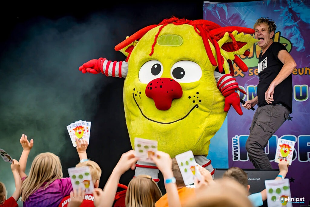 Kinder tanzen mit großem gelben Maskottchen auf Bühne.
