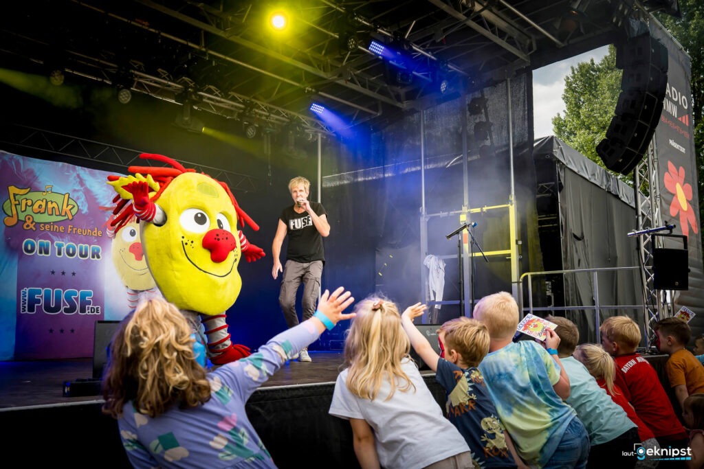 Kinder feiern bei Konzert mit Maskottchen auf der Bühne.