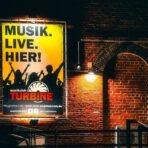 Plakat mit Musik-Event-Werbung an Backsteinwand.