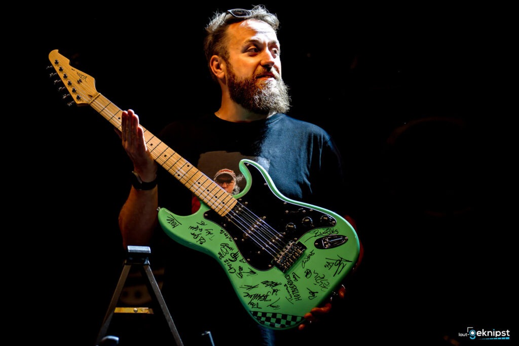 Mann hält signierte grüne E-Gitarre auf Bühne.