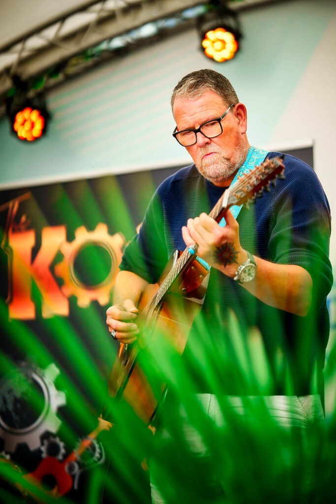 Mann spielt Gitarre auf Bühne mit Pflanzen im Vordergrund.