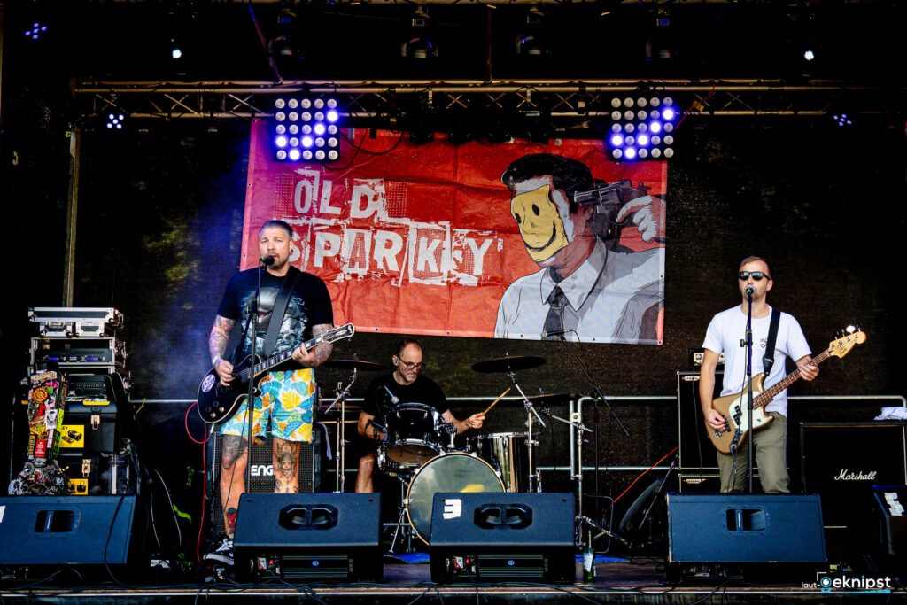 Band spielt auf Open-Air-Bühne mit Plakat im Hintergrund.