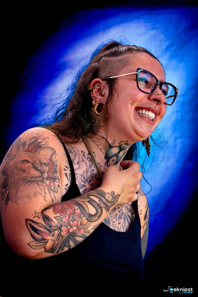 Lächelnde Person mit Tattoos und Piercings vor blauem Hintergrund