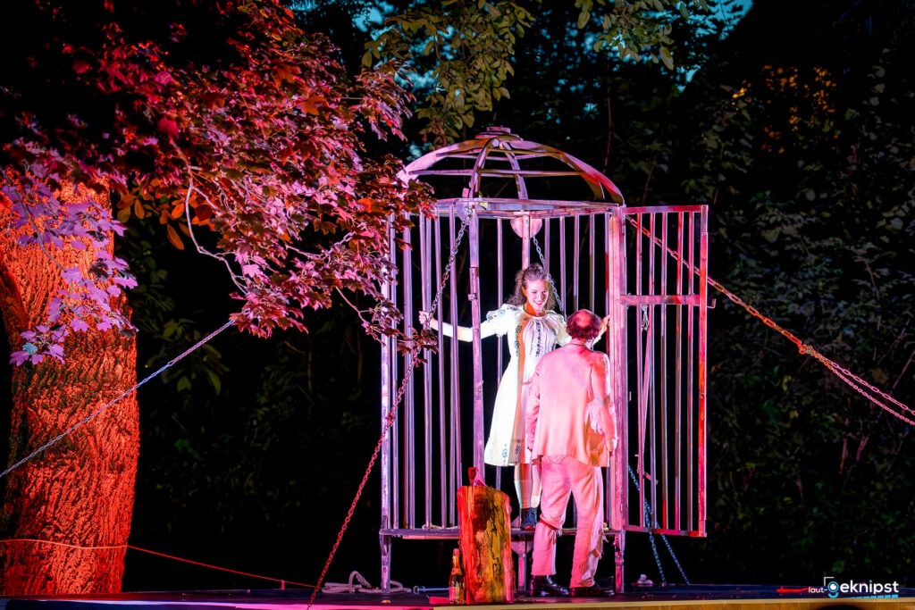 Frau im Käfig auf einer Waldtheaterbühne bei Nacht.