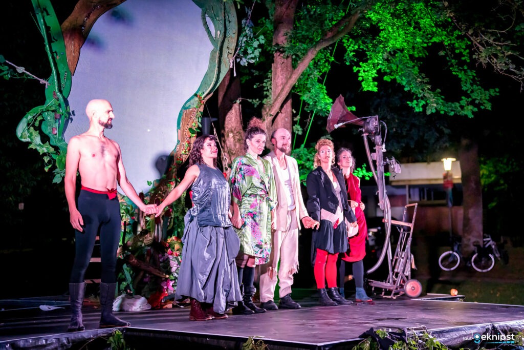 Theatergruppe verbeugt sich auf Außenbühne unter Bäumen.