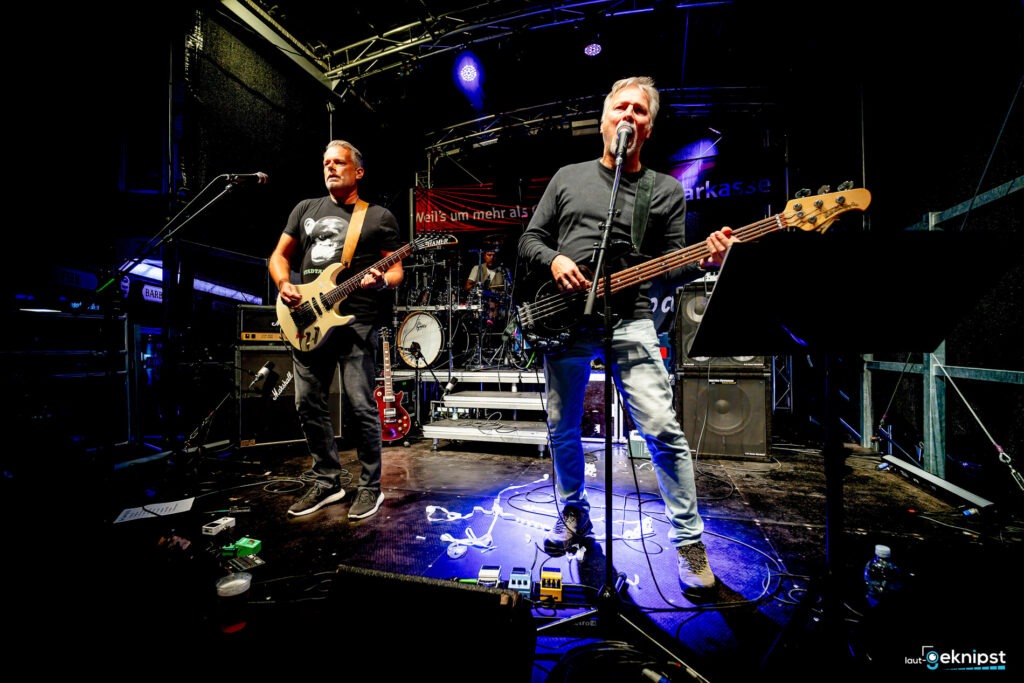 Band spielt auf Bühne, Gitarren, Mikrofone, Schlagzeug.