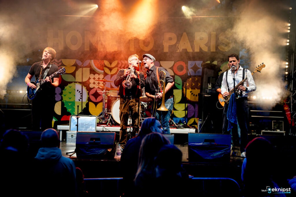 Band spielt live auf bunter Bühne vor Publikum.