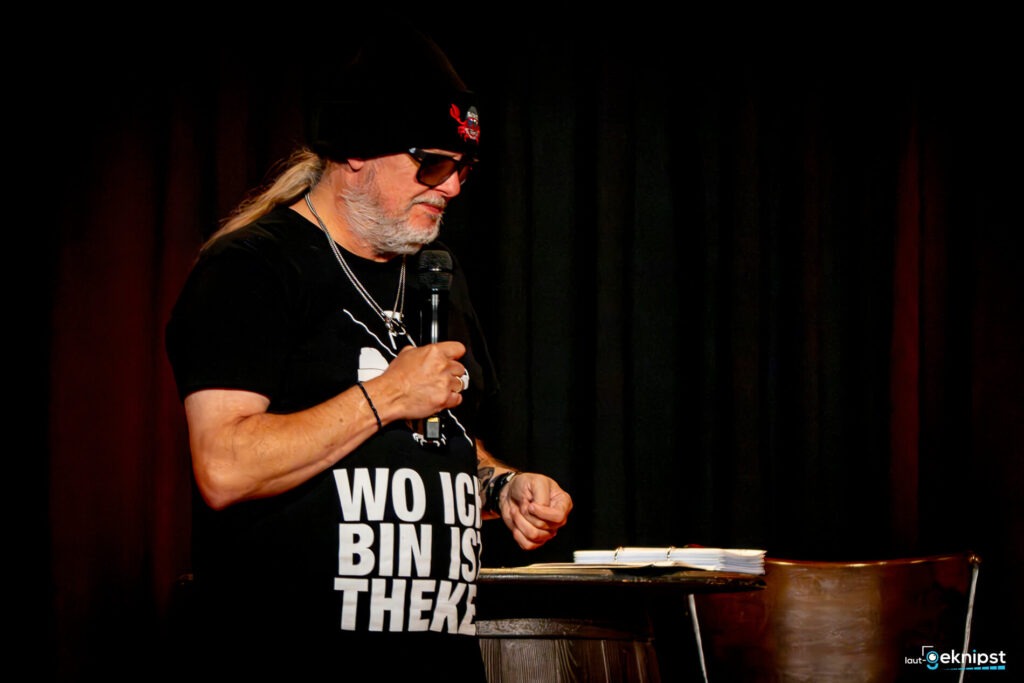Mann mit Mikrofon auf der Bühne, T-Shirt-Spruch sichtbar.