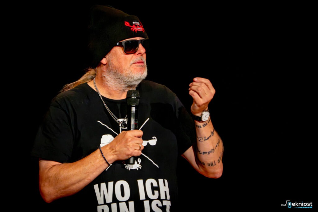 Person hält Mikrofon und trägt Mütze und Sonnenbrille.