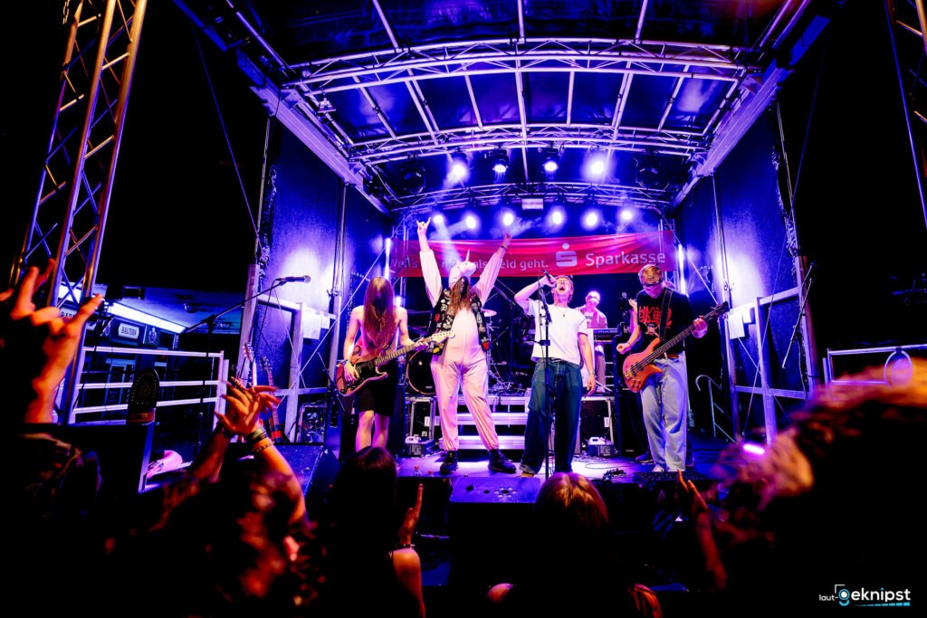 Band spielt auf beleuchteter Bühne, Publikum jubelt.
