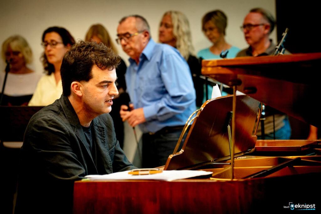 Mann spielt Klavier mit Chor im Hintergrund.