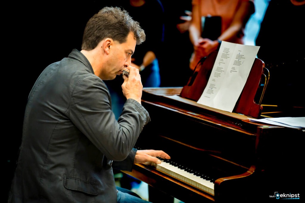 Musiker spielt Mundharmonika und Klavier gleichzeitig.