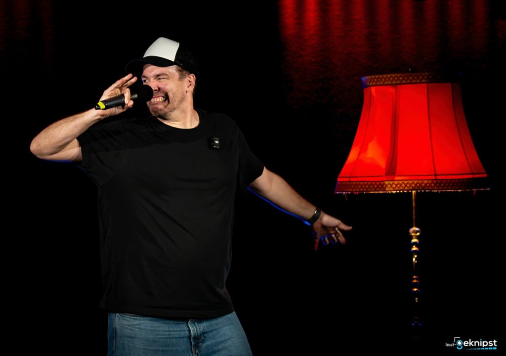 Mann singt auf Bühne neben roter Lampe.