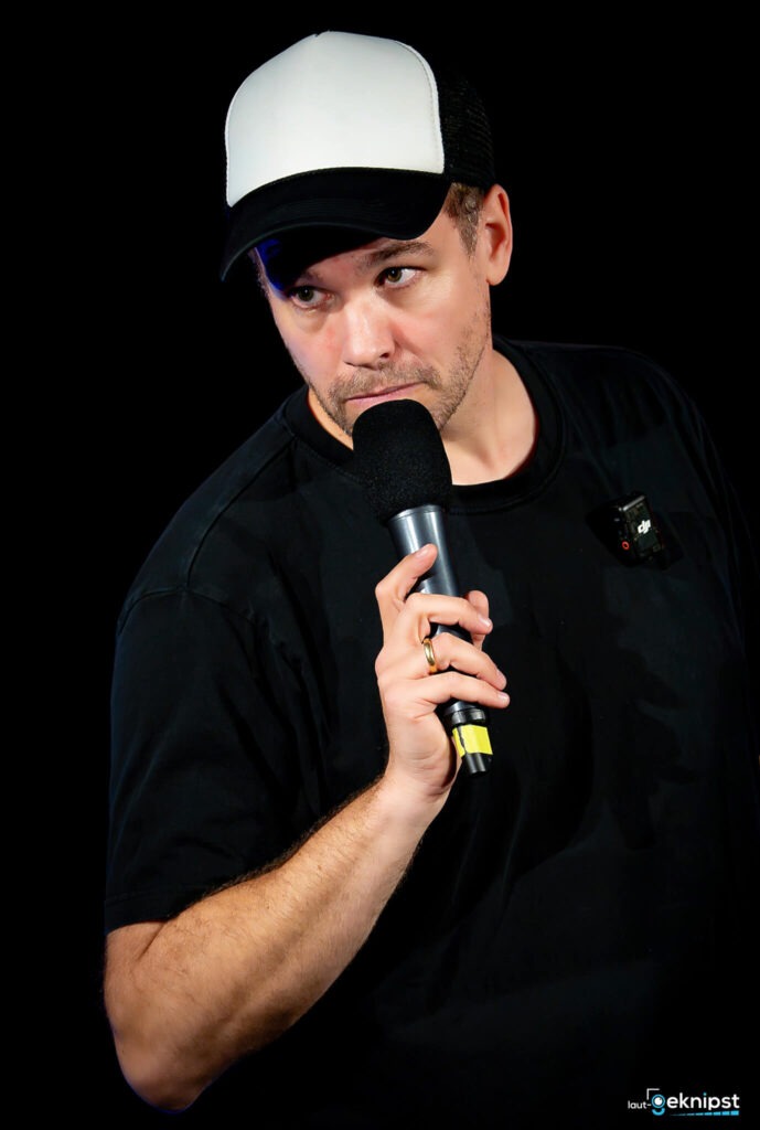 Person mit Mikrofon und Kappe, schwarzer Hintergrund.