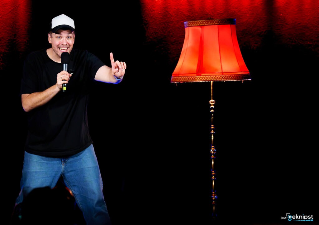 Komiker auf Bühne mit Mikrofon und Stehlampe.