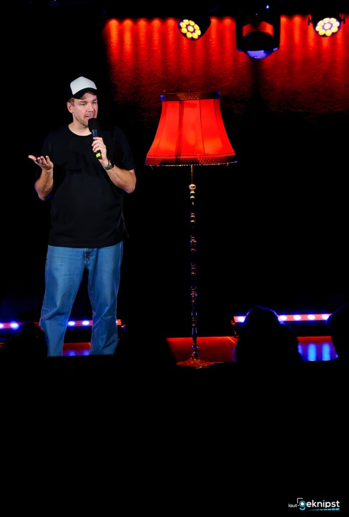 Stand-up-Comedian spricht neben roter Lampe auf Bühne.